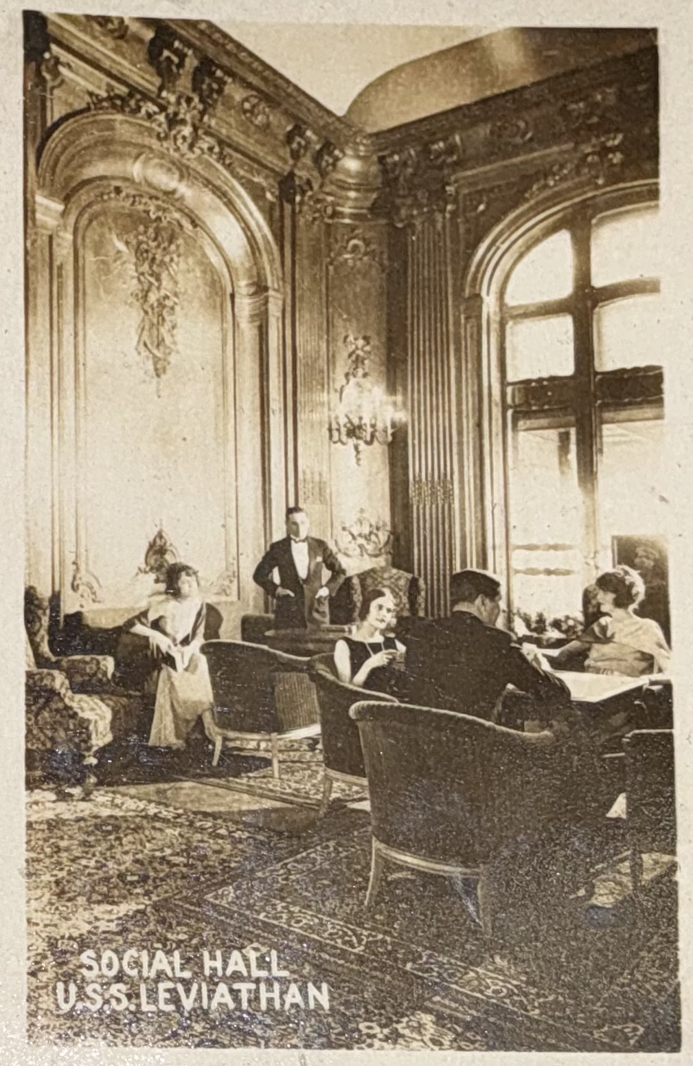 Passengers seated in the social hall with tall windows.